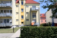 Gelbes Haus mit angebauten Balkonen, im Hintergrund orangenes Haus, vorne niedrige Hecken.