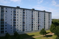 Sanierter Wohnblock in hellen Blautönen, Brüstungen Loggien mittelblau. Flachdach, 9 Etagen. Davor Grünfläche mit Weg und Bäumen.