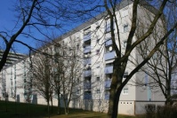 Sanierter, blaugrauer Wohnblock. Flachdach. Brüstungen Loggien in mittelblau. Davor hohe Bäume im Winter.