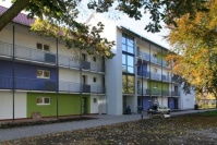 Wohnhaus mit Laubengängen, links drei Etagen, rechts vier Etagen. Vorne Blätter von einem Baum.