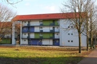 Weißes Wohnhaus mit drei Etagen. Auf den Laubengängen farbliche Akzente in grün und blau. Wiese mit Bäumen.