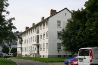 Mehrfamilienhaus mit Satteldach, 2 Hauseingängen, 3 Etagen. Weiße Fassade. Rechts hohe Bäume und 3 parkende Autos.
