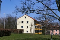 Giebelseite saniertes, gelbes Mehrfamilienhaus, Satteldach, 3 Etagen. Vorne Grünfläche mit niedrigen Hecken, Bäume im Winter. 