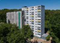 Hochhaus mit 2 Baukörpern, linker Teil mit Baugerüst und Netzen, rechter Teil weiße Fassade mit bunten Balkonbrüstungen. Rundum Bäume.