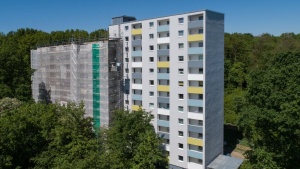 Hochhaus mit 2 Baukörpern, linker Teil mit Baugerüst und Netzen, rechter Teil weiße Fassade mit bunten Balkonbrüstungen. Rundum Bäume.