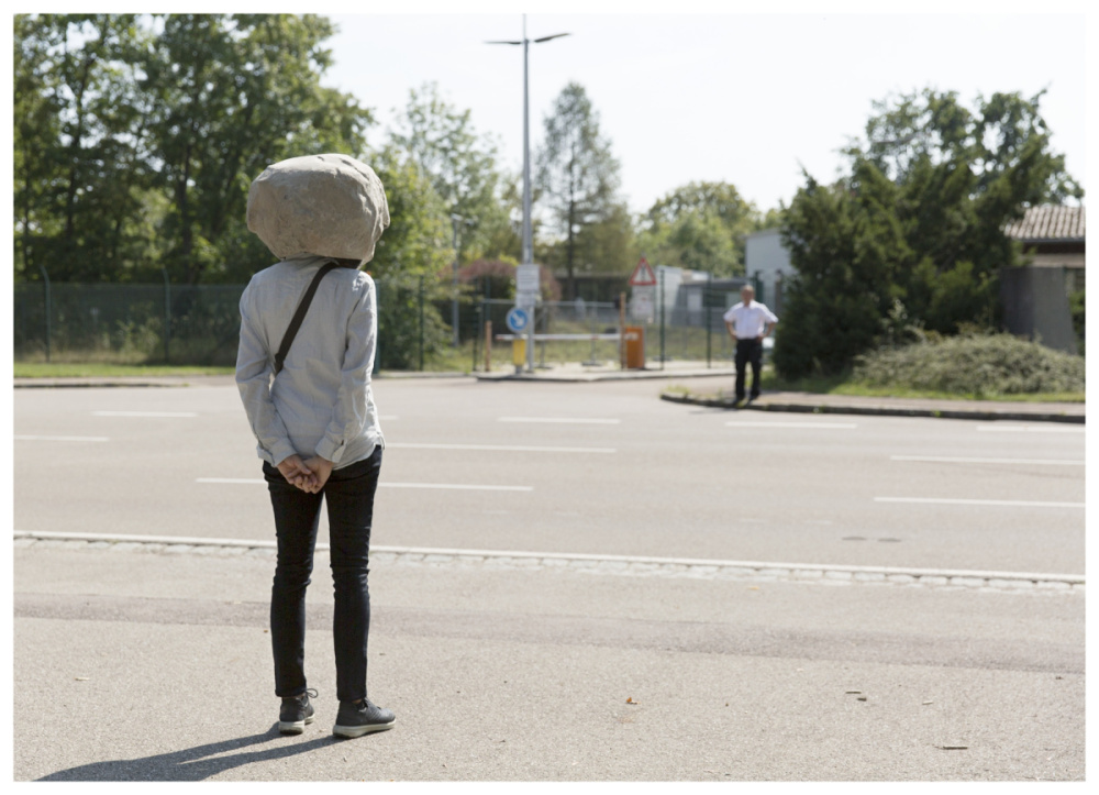 Person wears a stone as a head and stands on a wide road