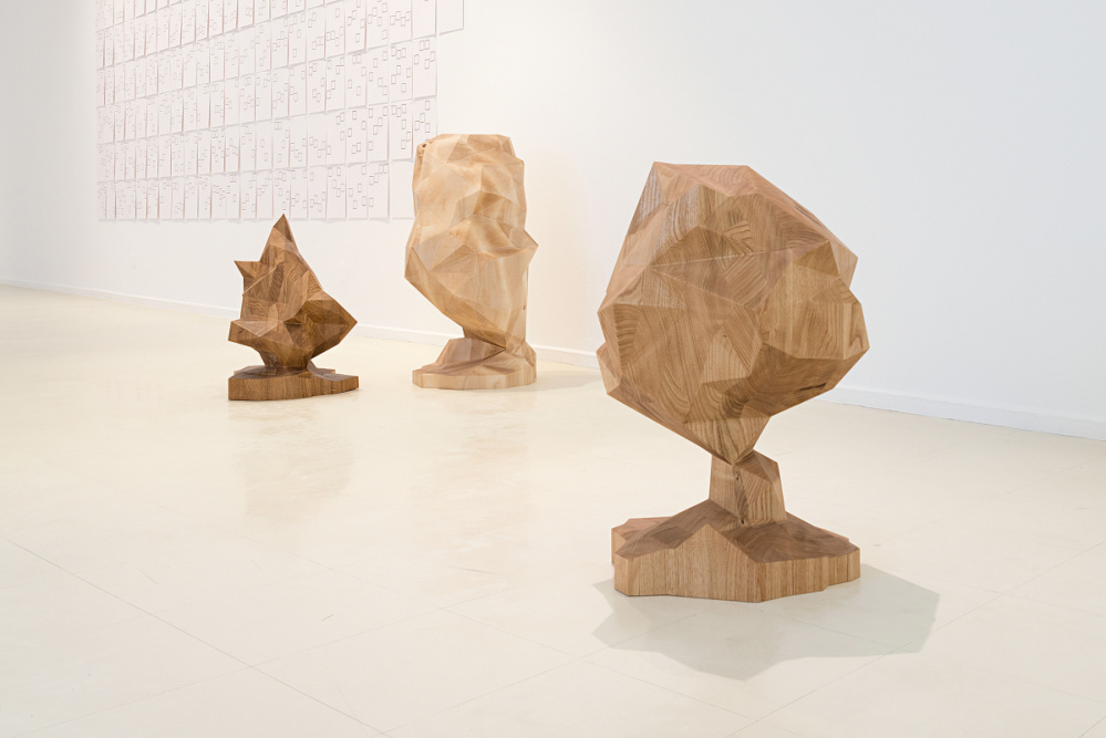 View into an exhibition space. Three sculptures milled from wood, reminding of scaled-down trees. In the background, DINA4 papers in rows with drawings attached to a wall.