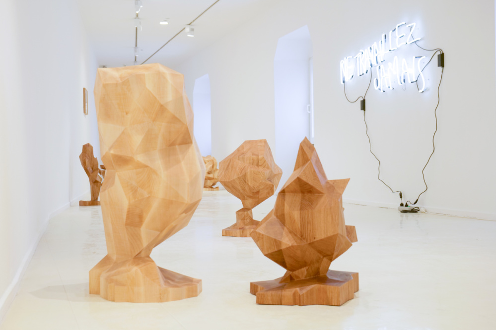 View into an exhibition space.Sculptures milled from wood, reminding of scaled-down trees. In the background a neon sign "Ne travaillez jamais."