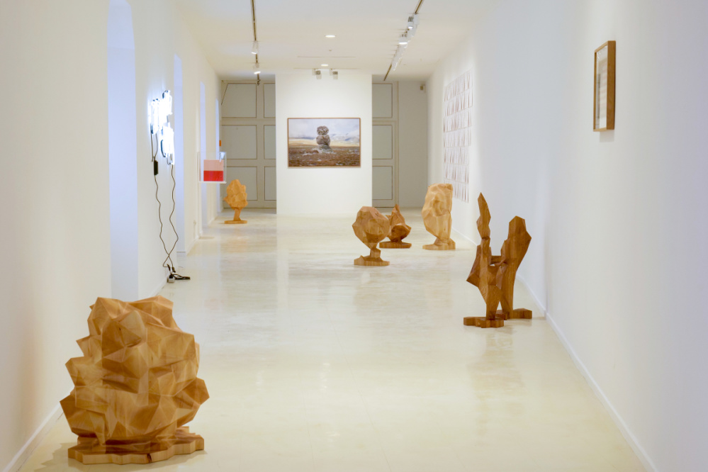 View into an exhibition space, freely distributed, sculptures milled from wood, reminiscent of scaled-down trees. In the background, a large photo of three stones stacked on top of each other. DINA4 papers in rows with drawings mounted on a wall.