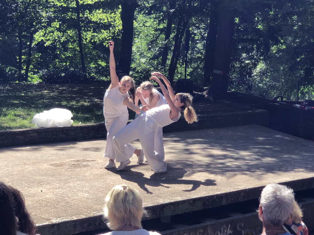 Drei Frauen in weißer Kleidung tanzen auf einer Bühne im Wald