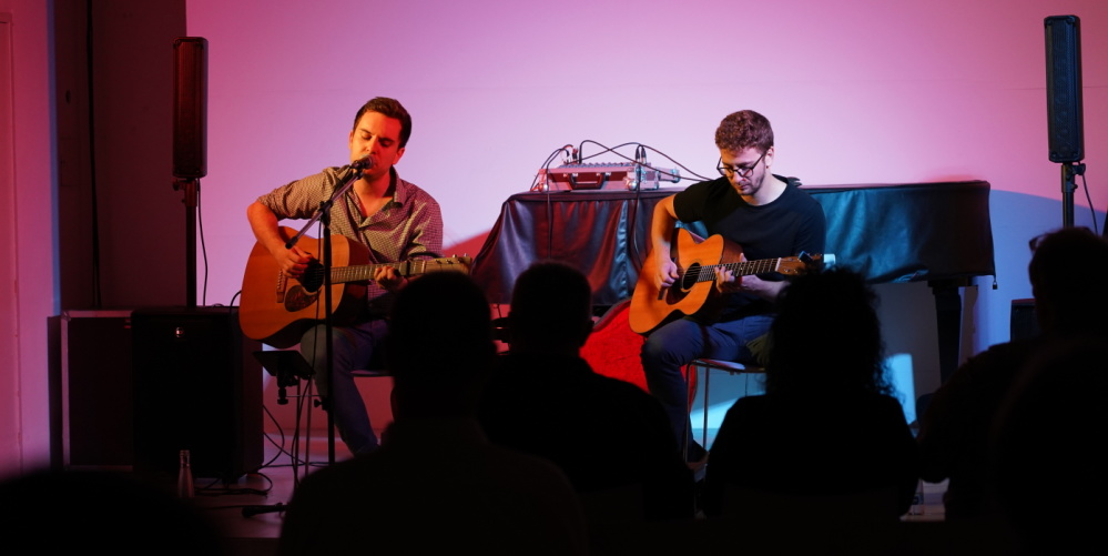 Veranstaltungsraum der Stadtgalerie, Umrisse von einem Publikum, zwei männliche Gitarrenspieler auf der Bühne