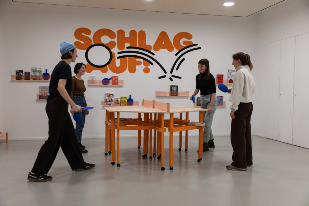 Vier Personen, die Tischtennis spielen. Im Hintergrund eine weiße Wand mit dem orangen Schriftzug Schlag Auf! und Regale mit Büchern