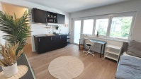 Wohnraum mit schwarzer Miniküche, Schreibtisch mit Stuhl am Fenster, Regal und Einzelbett