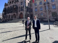 Zwei Personen in Anzügen stehen vor einem Fahnenmast, im Hintergrund befindt sich das Rathaus St. Johann