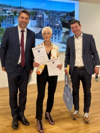 Links  steht ein Mann im Anzug, in der Mitte eine Frau mit zwei Urkunden und Tüte, rechts ein Mann mit einer Tüte und Mikrofon