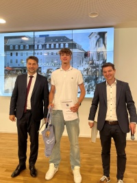 Links  steht ein Mann im Anzug mit Mikrofon, in der Mitte ein junger großer Mann mit Urkunde und Tüte, rechts ein Mann mit einem Umschlag und Mikrofon