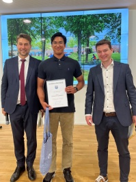 Links  steht ein Mann im Anzug mit Mikrofon, in der Mitte ein Mann mit Urkunde und Tüte, rechts ein Mann mit einem Umschlag