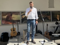 Ein Mann mit Mikrofon steht auf einer kleinen Bühne. Im Hintergrund sieht man Musikinstrumente