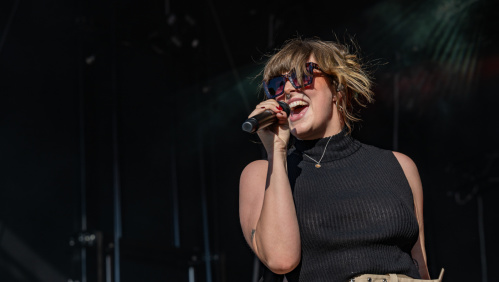 Sophie Sängerin singt leidenschaftlich mit Sonnenbrille und schwarzem Top vor dunklem Bühnenhintergrund.