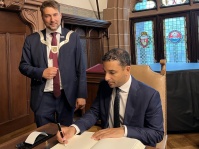 Ein Mann sitzt schreibend auf einem Stuhl, ein anderer steht neben ihm