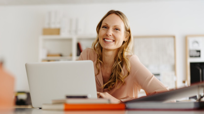 Überweisungen und Daueraufträge Eine lachende blonde Frau sitzt an einem Schreibtisch vor einem aufgeschlagenen Laptop. Foto: contrastwerkstatt / AdobeStock