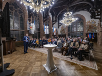 Ein Mann steht in einem festlichen Saal vor Publikum und erklärt diesem etwas