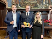 Zwei Männer und eine Frau in Anzügen und stehen in einem historischen Saal nebeneinander und posieren für die Kamera. Der Mann in der Mitte hat einen Miniaturlöwen in der Hand