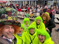 Mehrere Tänzerinnen in grün-gelben Kostümen mit großen weiß-gelben Perücken führen eine Choreografie auf der Straße auf.