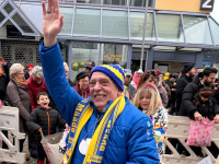 Ein Mann in blauer Jacke, blau-gelber Mütze und Schal winkt lachend in die Kamera, hinter ihm stehen viele Zuschauer am Straßenrand.